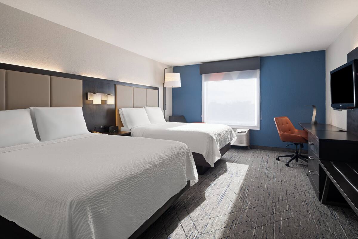 A hotel room with two beds, a desk, a chair, a TV, and a large window.