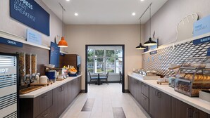 A modern breakfast area with a counter displaying cereals, a bakery section, and a seating area with a view through a large window. - Holiday Inn Express St. Petersburg North (I-275) by IHG (St. Petersburg)