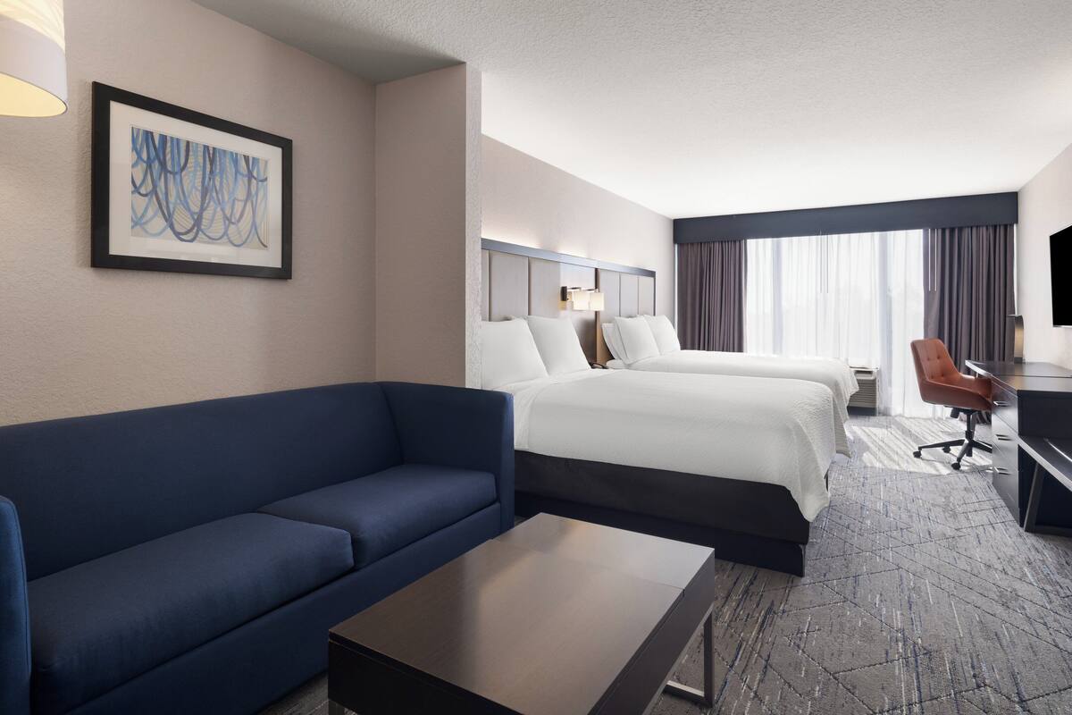 A hotel room with two beds, a blue sofa, a desk, and a large window with curtains.