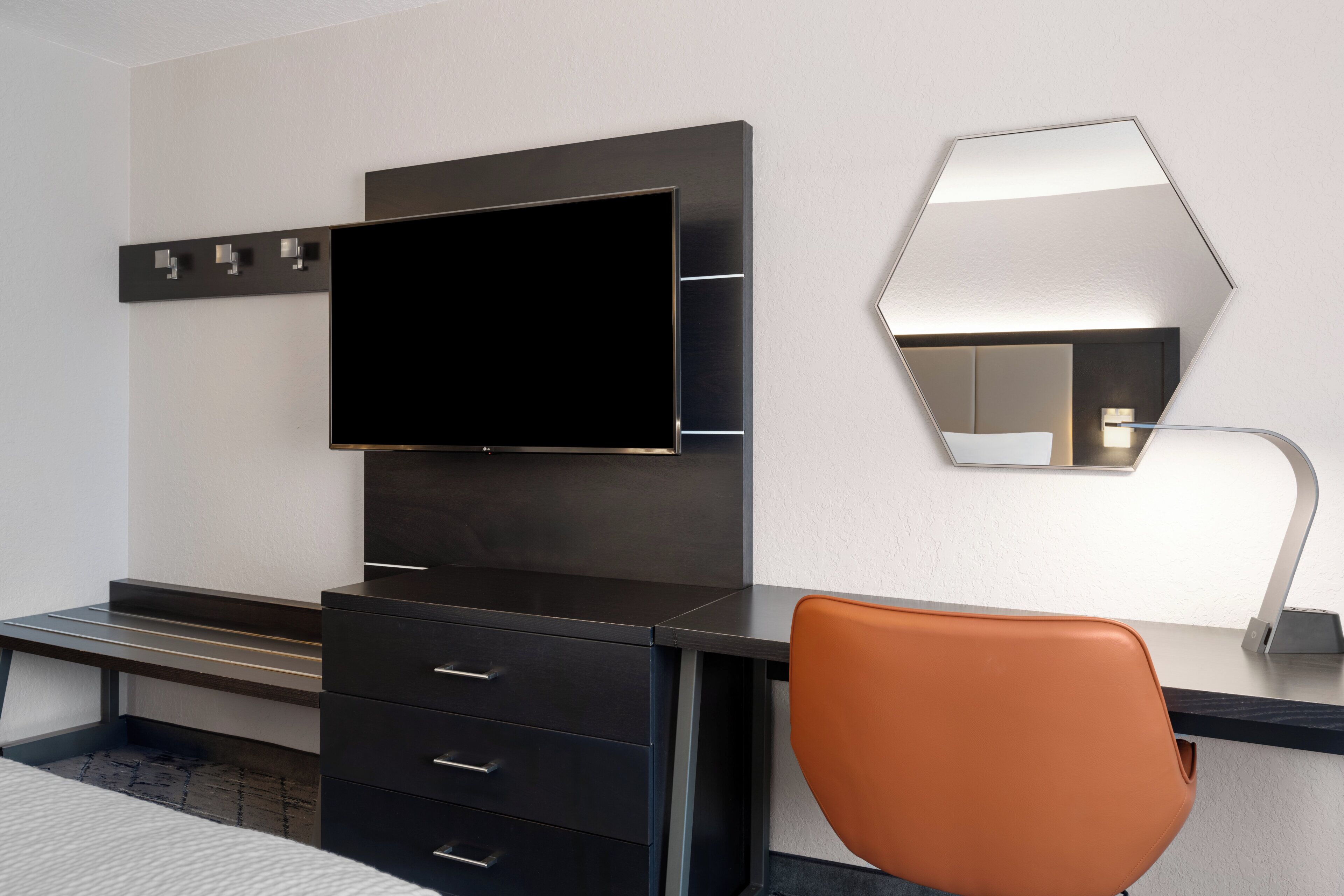Una habitación de hotel moderna con un televisor de pantalla plana montado en un mueble de madera oscura, un espejo grande en la pared y una sola silla naranja.
