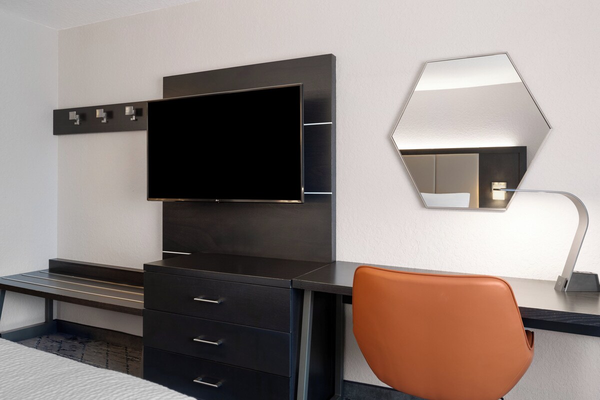 Quarto de hotel moderno com televisão de tela plana presa a um móvel de madeira escura, um espelho grande na parede e uma única cadeira laranja.