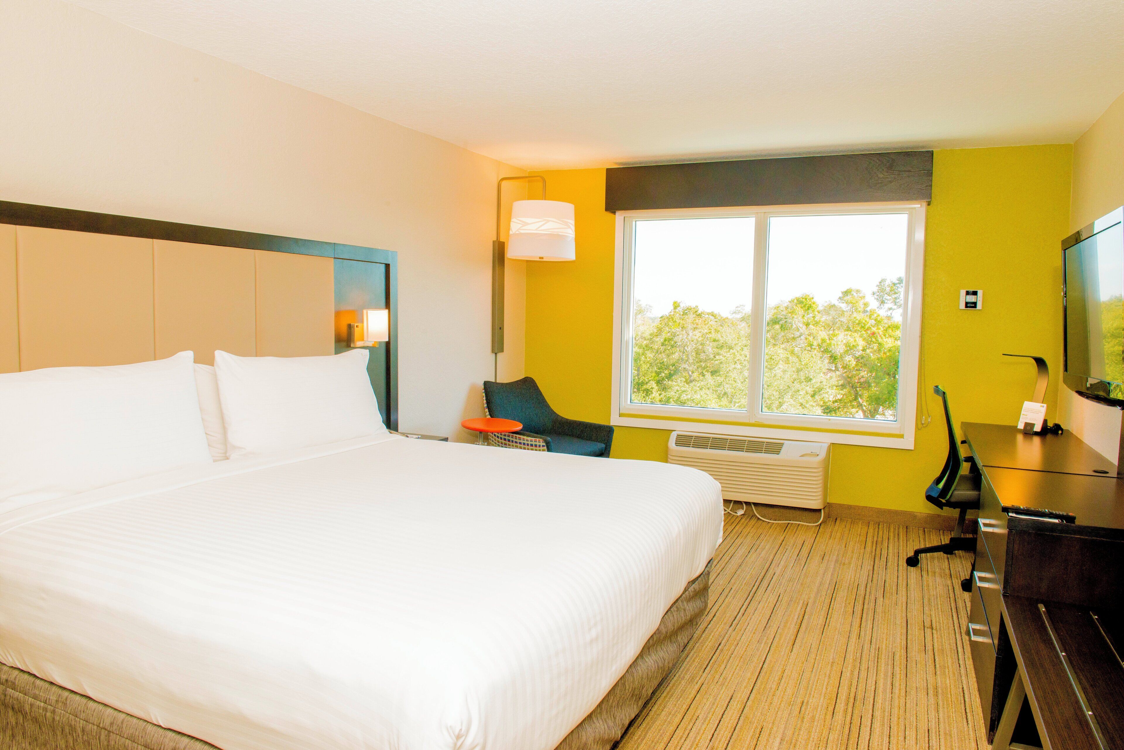 Habitación de hotel con una cama grande, un escritorio, una silla, un televisor y una ventana con vista a árboles.