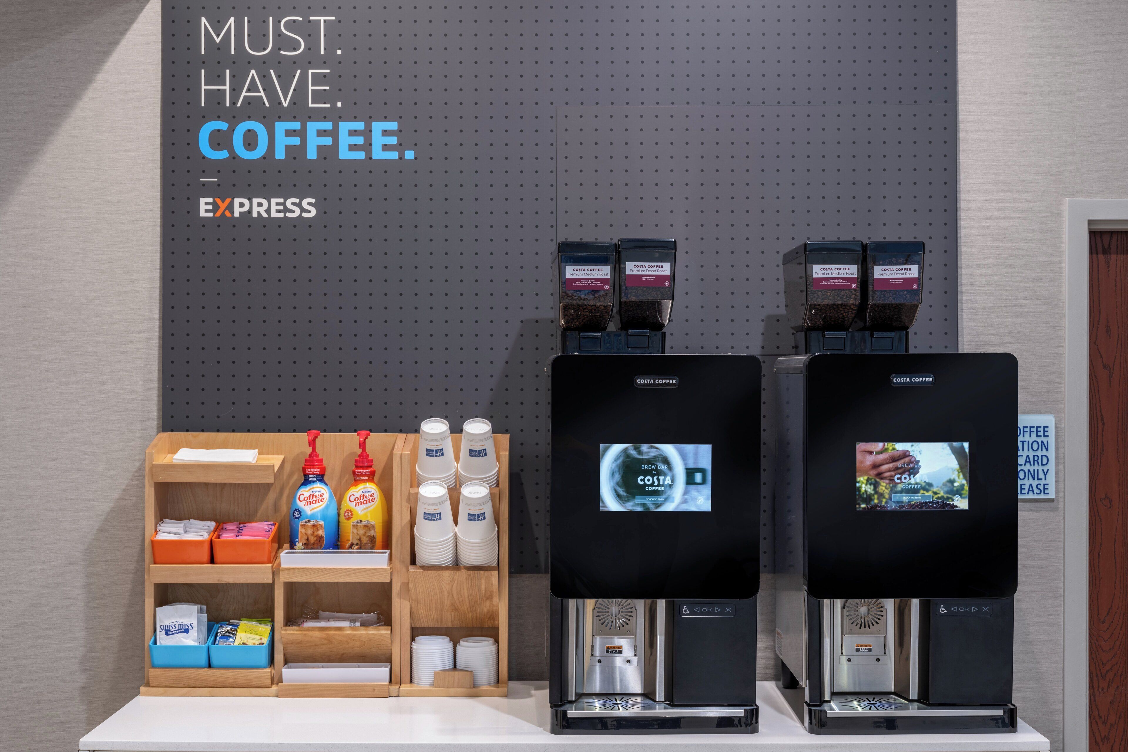 Uma estação de café com duas máquinas de café, identificadas como 'COSTA COFFEE', e uma seleção de cápsulas e xícaras.