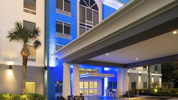 A hotel entrance with a blue-lit facade, a covered walkway, and a palm tree. - Holiday Inn Express St. Petersburg North (I-275) by IHG (St. Petersburg)