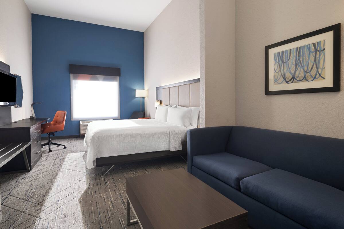 A hotel room with a bed, a blue sofa, a desk with a chair, a TV, and a painting on the wall.