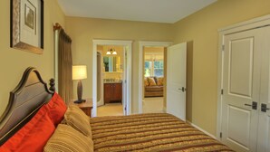 Une chambre d’hôtel avec un lit, une table de chevet, une lampe, une porte et une vue sur une autre chambre qui contient un canapé et une fenêtre.