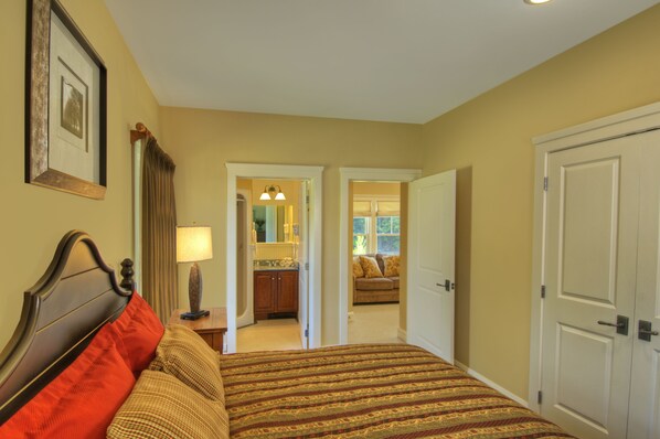 A hotel room with a bed, a nightstand, a lamp, a door, and a view into another room with a couch and a window.