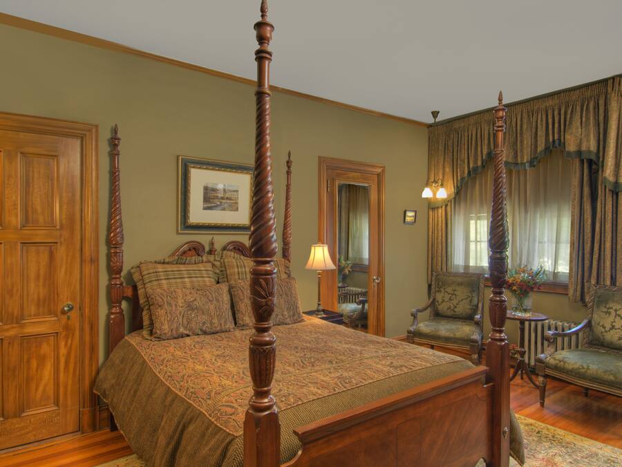 A traditional bedroom with a four-poster bed, two armchairs, a wooden wardrobe, and a mirror.