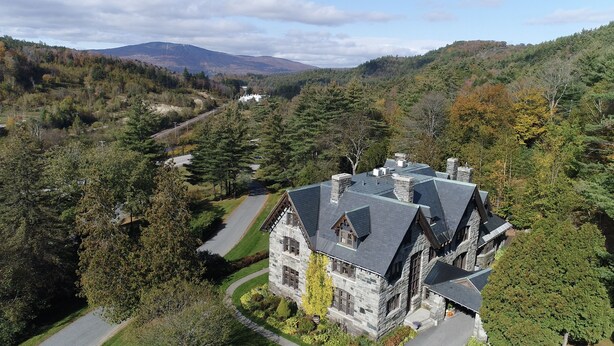 Un grand bâtiment en pierre, doté d’un toit à forte pente, entouré d’une forêt dense et de collines.