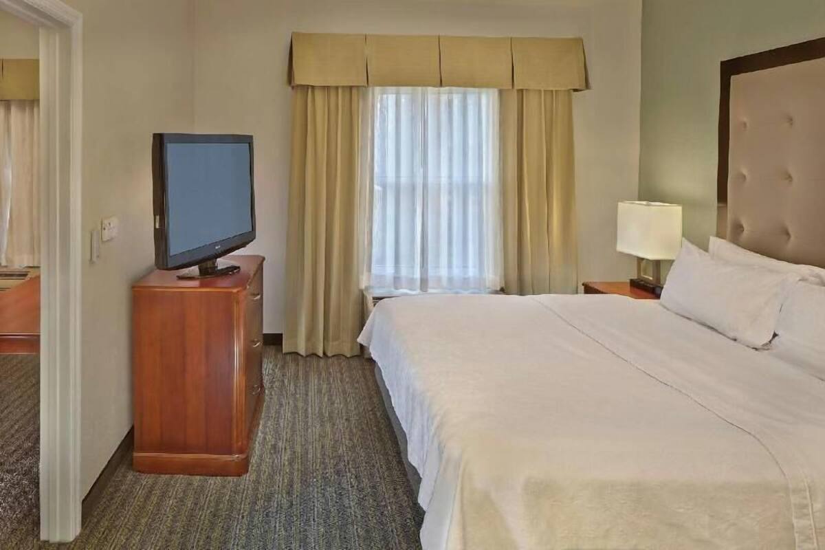 A hotel room with a bed, a television on a wooden stand, a window with curtains, and a bedside lamp.
