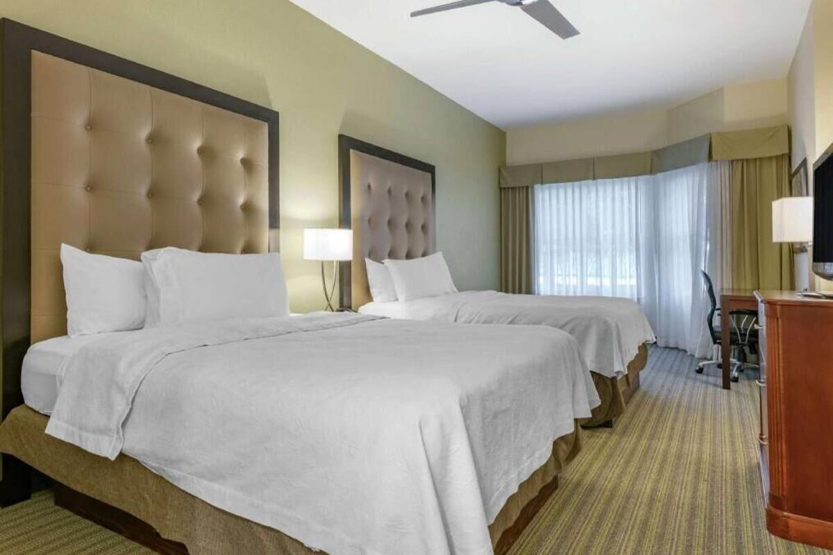 A hotel room with two beds, a desk, a chair, a television, and a ceiling fan.