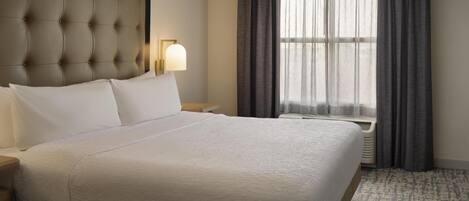 A hotel room with a large bed, a headboard, a bedside lamp, a window with curtains, and a carpeted floor.