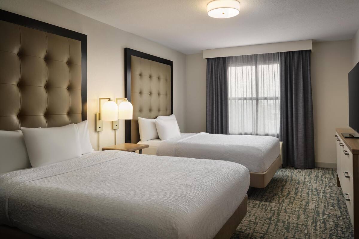 A hotel room with two beds, a large tufted headboard, a bedside lamp, a large window with curtains, and a wooden dresser.