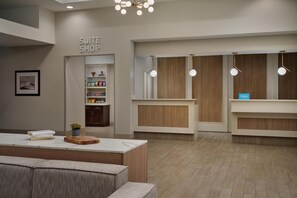 Un espace d’accueil d’hôtel avec une enseigne indiquant « SUITE SHOP », un comptoir de réception, un coin salon avec un canapé et une étagère murale contenant des livres et une petite plante.