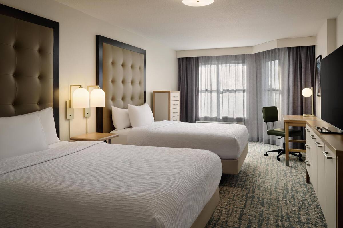 A hotel room with two beds, a desk, a chair, a TV, and a large window with curtains.