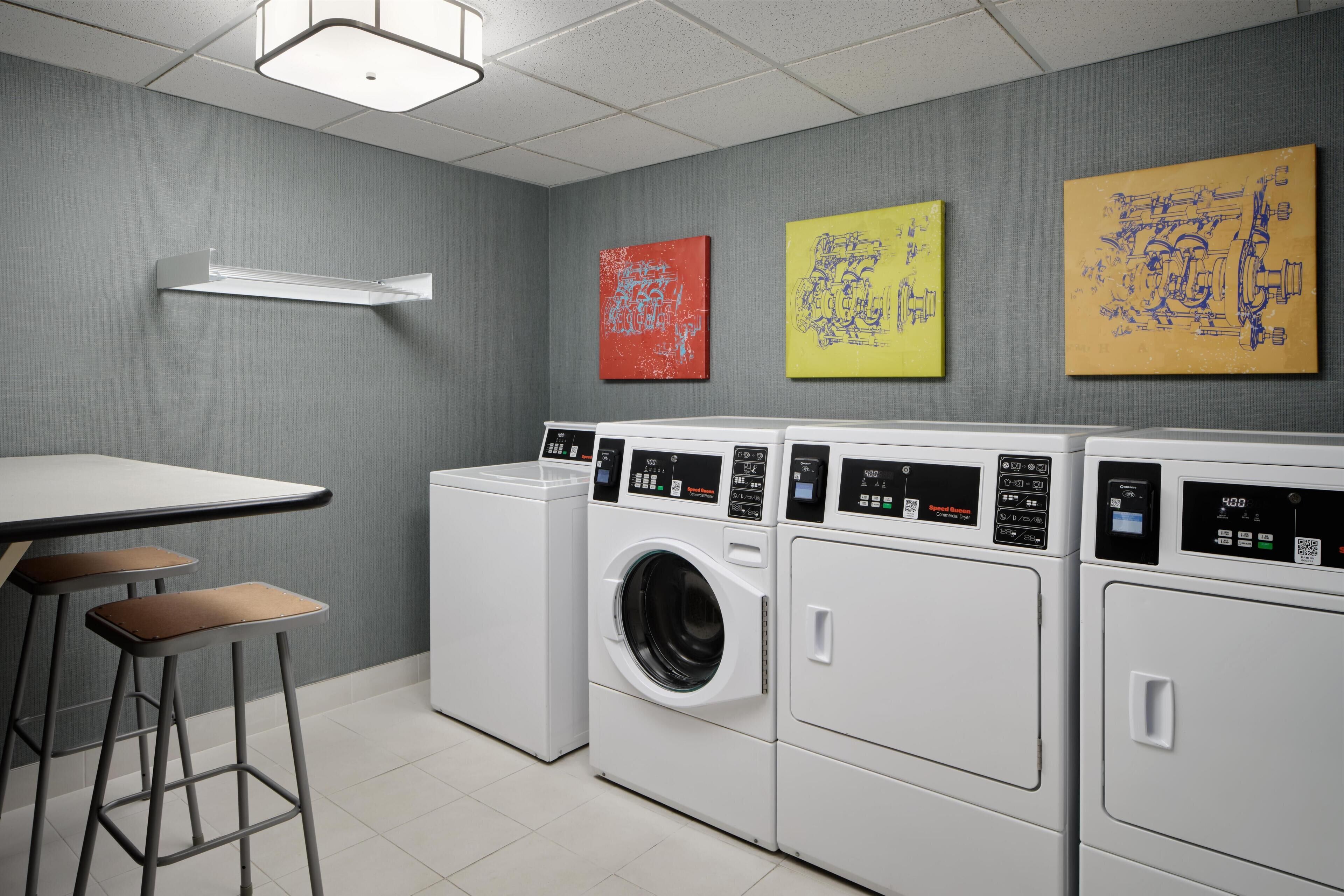 A laundry room with a table and stools, a washing machine, and three artwork pieces on the wall.