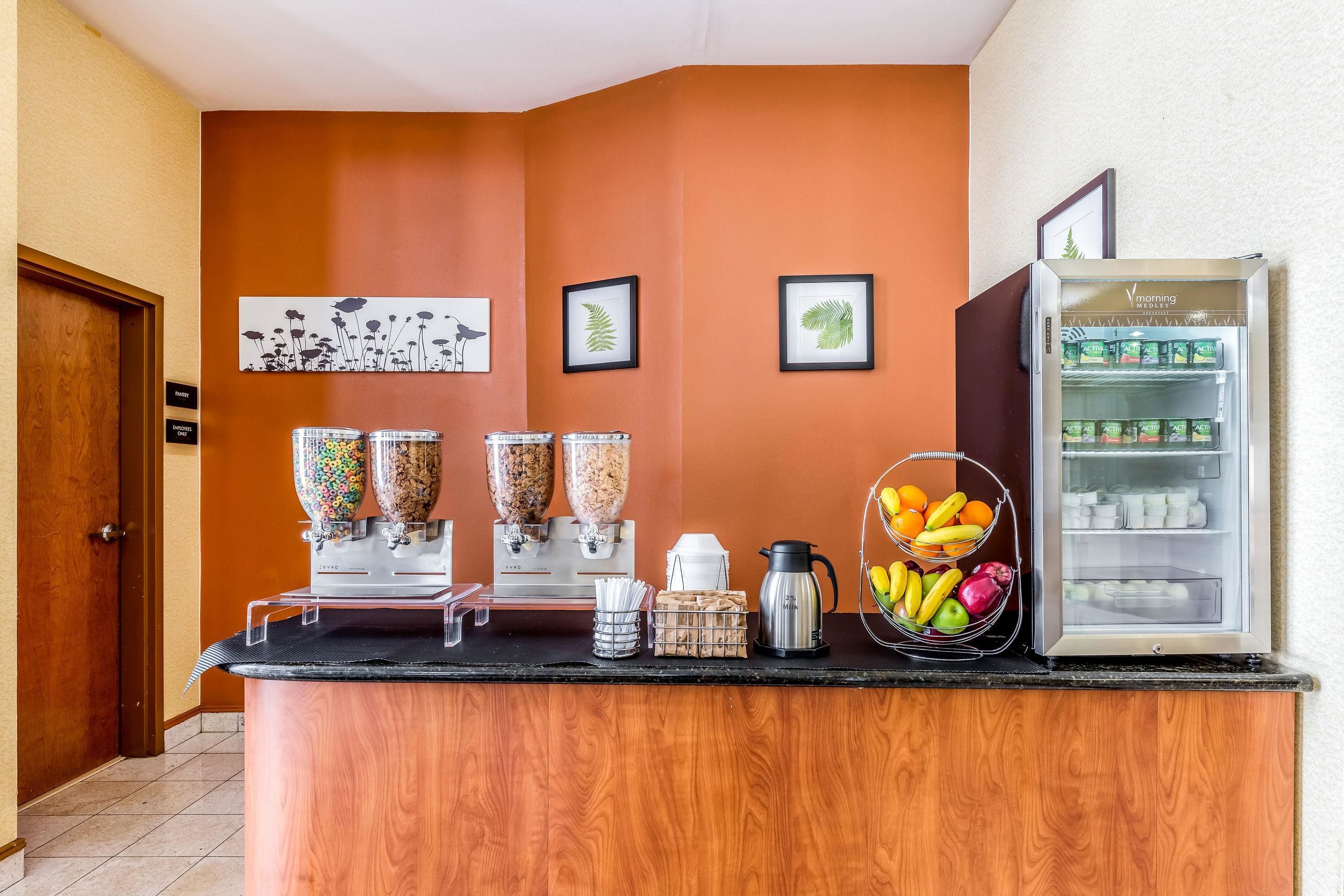 A hotel reception area with a wooden counter, a coffee machine, a fruit basket, a beverage dispenser, and a refrigerator stocked with drinks.