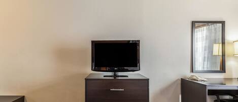Habitación de hotel con un aparador de madera oscura, un televisor de pantalla plana encima, un espejo sobre el aparador y un escritorio con lámpara y teléfono.