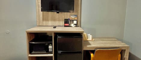 A compact hotel room with a wooden desk, a television, a refrigerator, and a single chair.