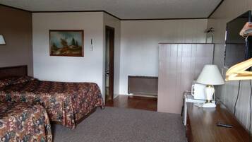 A hotel room with two beds, a desk with a lamp, a radiator, and a painting on the wall.