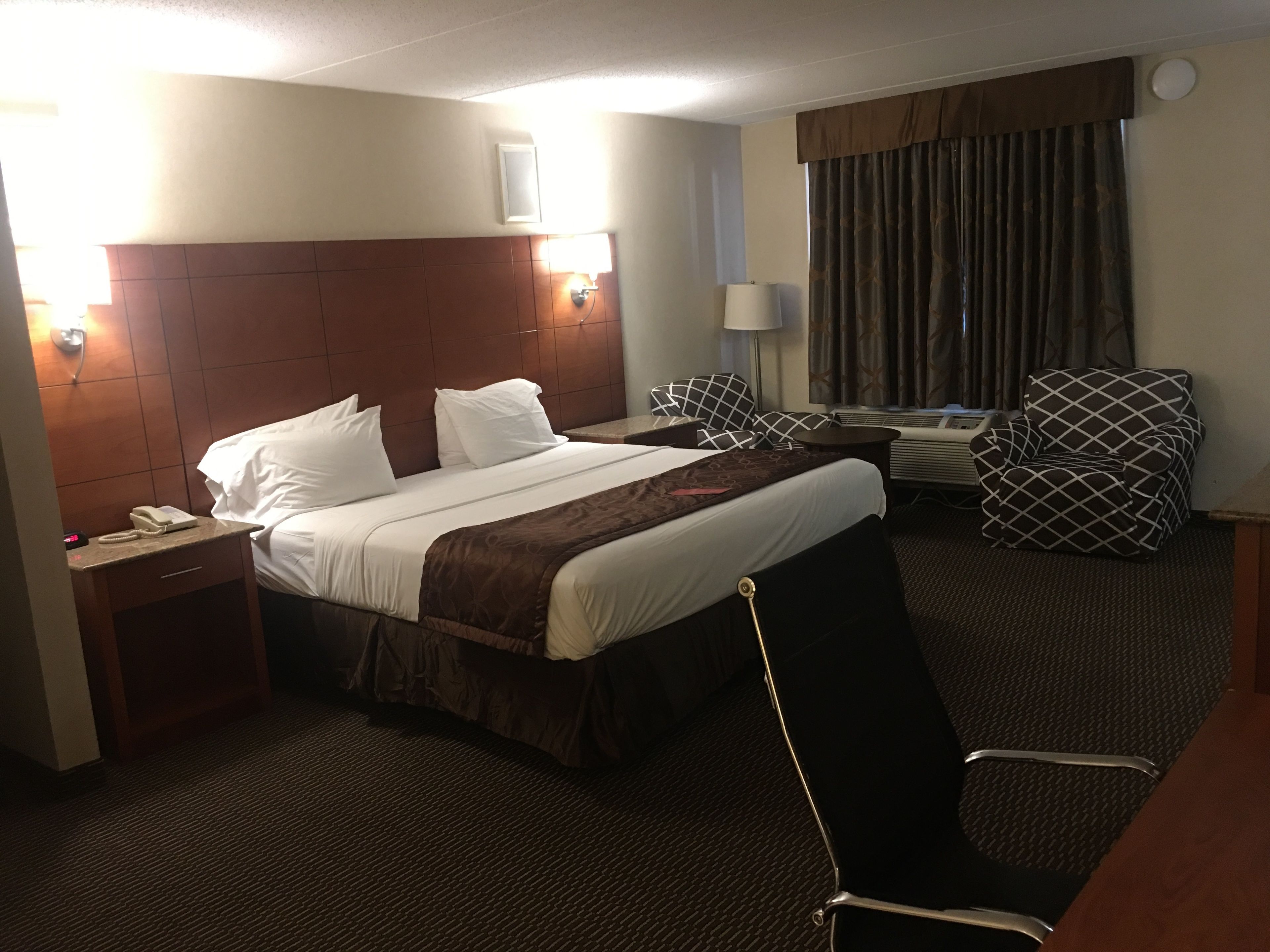 A hotel room with a large bed, a bedside table with a phone, a chair, and a desk with a lamp.