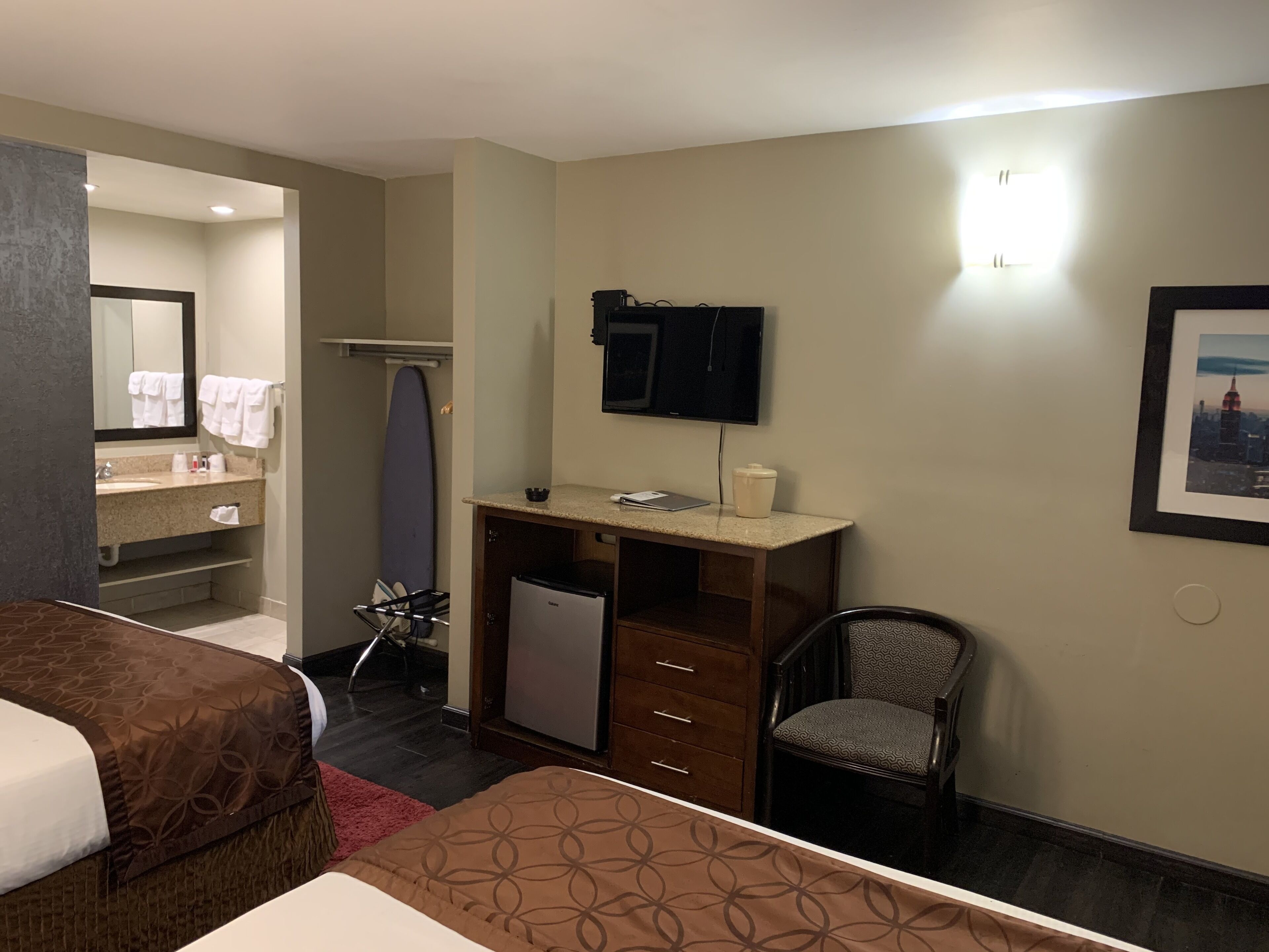 A hotel room with a bed, a desk with a computer, a chair, a small refrigerator, a TV, and a bathroom with a sink and mirror.