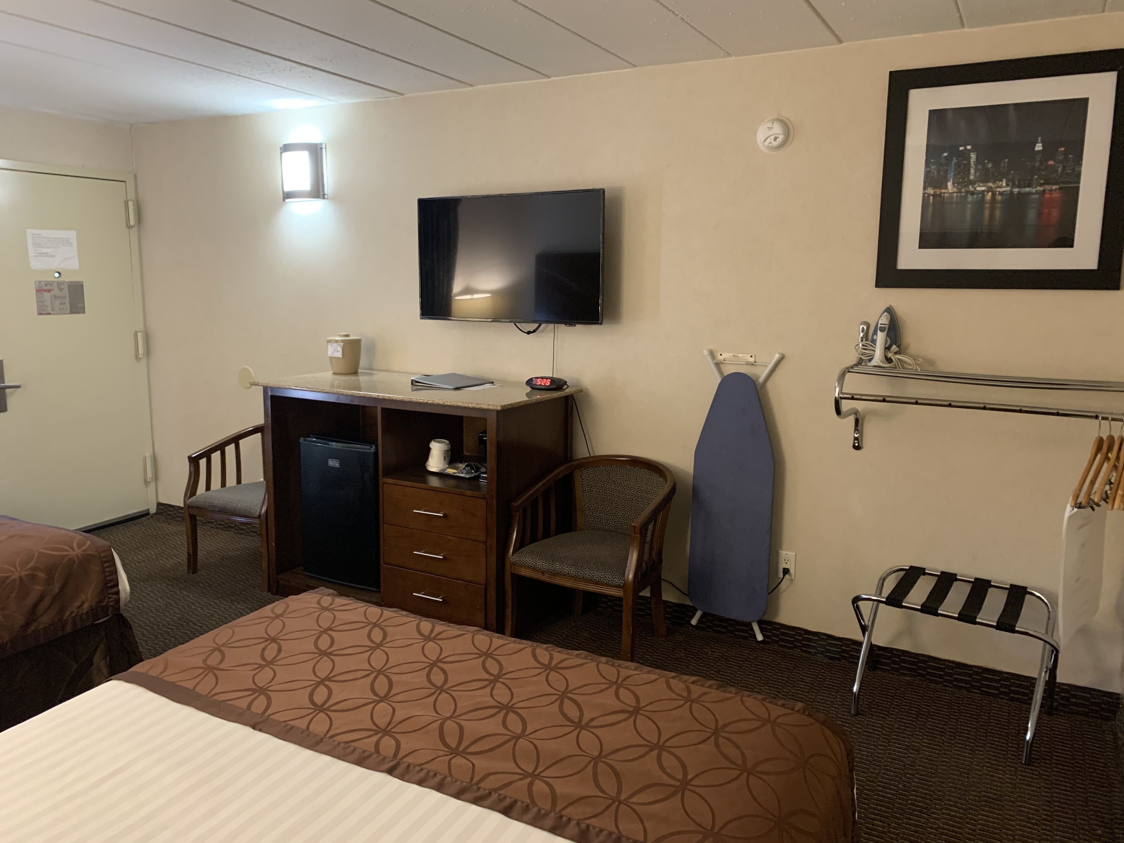 A hotel room with a bed, a desk with a computer, a chair, a TV, an ironing board, and a rack with clothes.