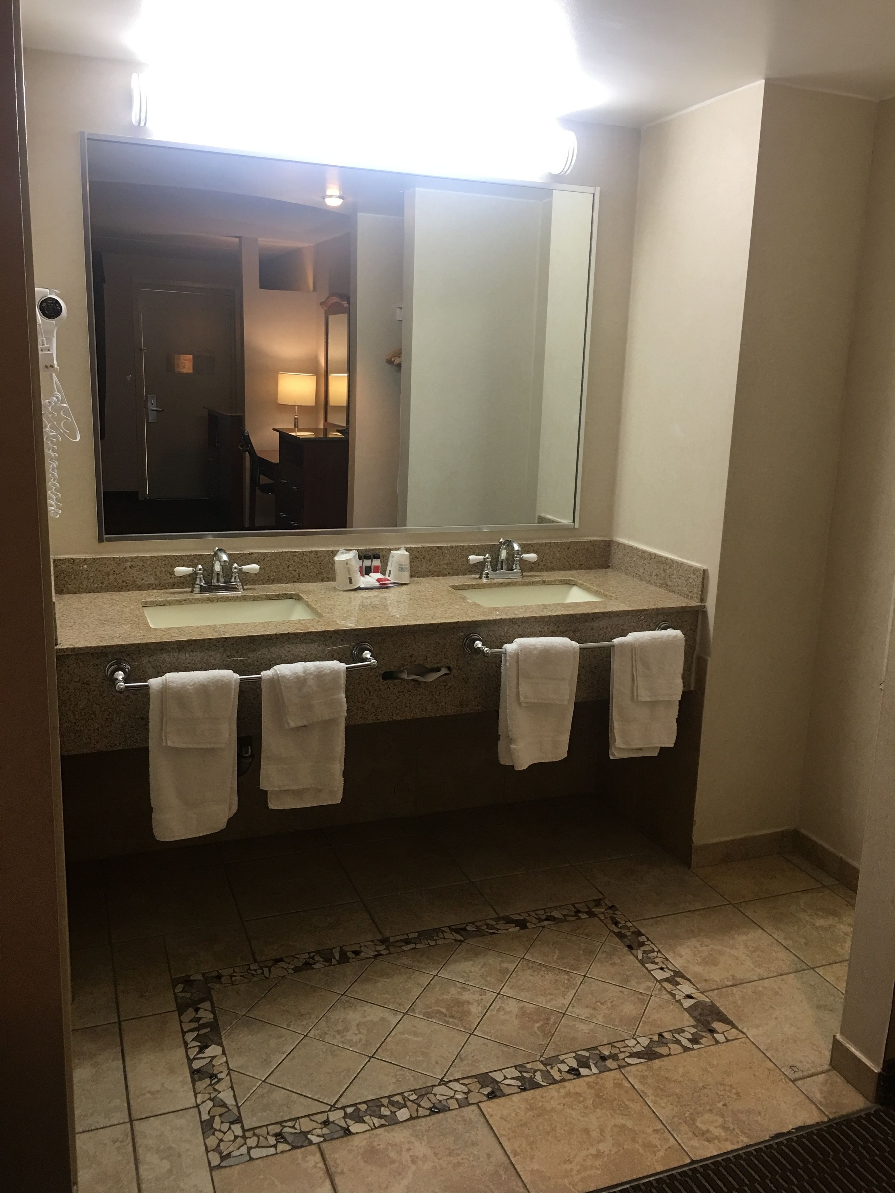 Une salle de bain avec deux lavabos, un grand miroir et un sol carrelé agrémenté d’une bordure décorative.