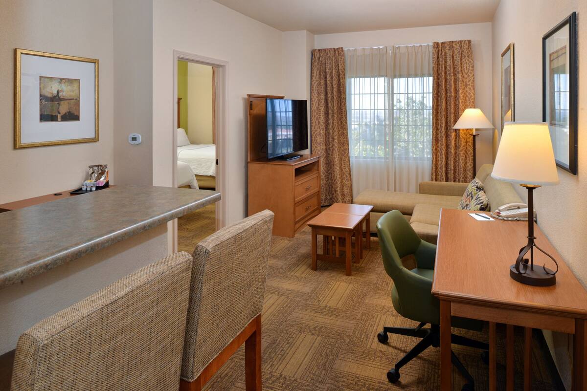 A hotel room with a kitchenette, a television, a sofa, a desk with a lamp, and a window with curtains.