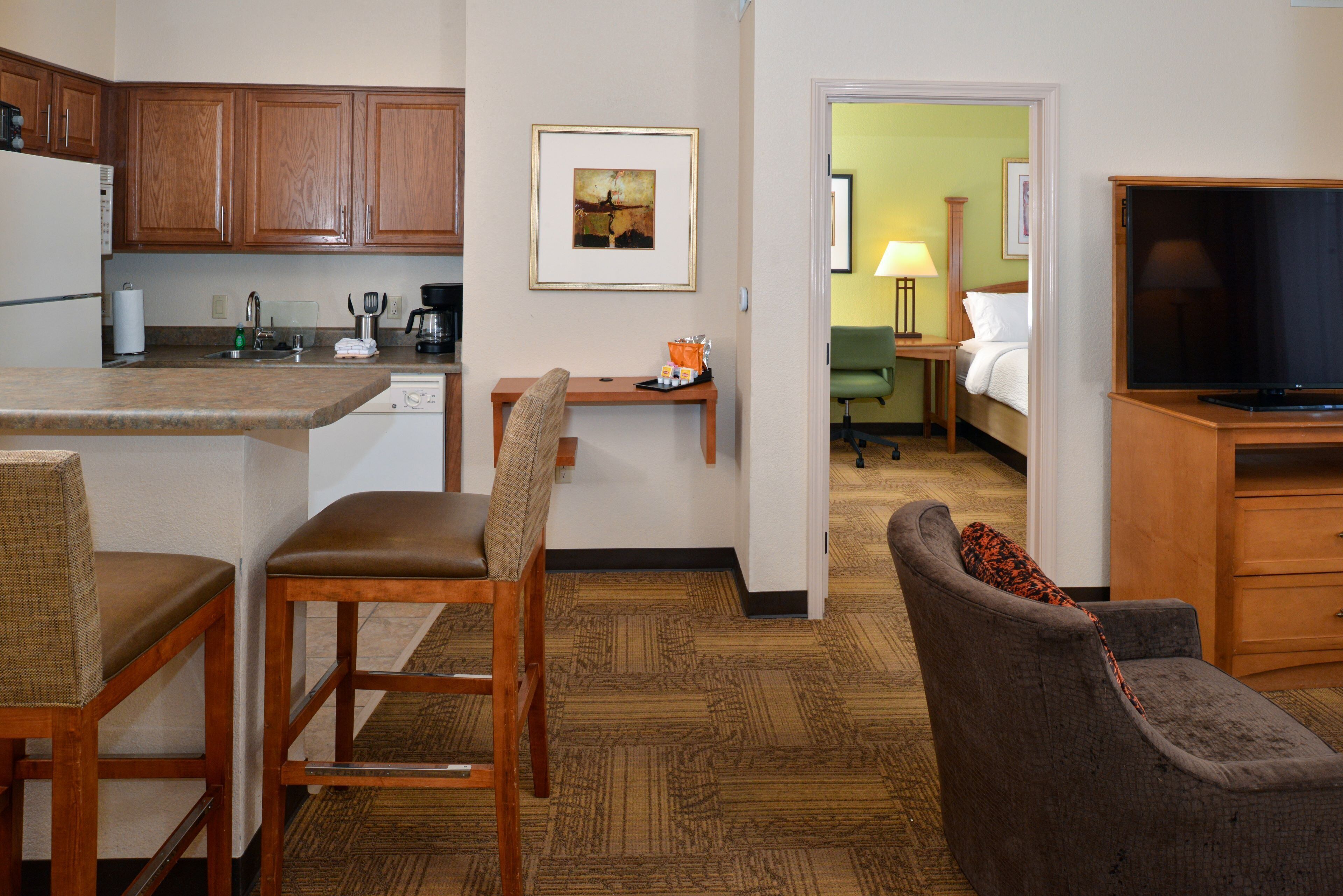 A hotel room with a kitchen area, a dining table with chairs, a sofa, a television, and a bed with a green wall in the background.