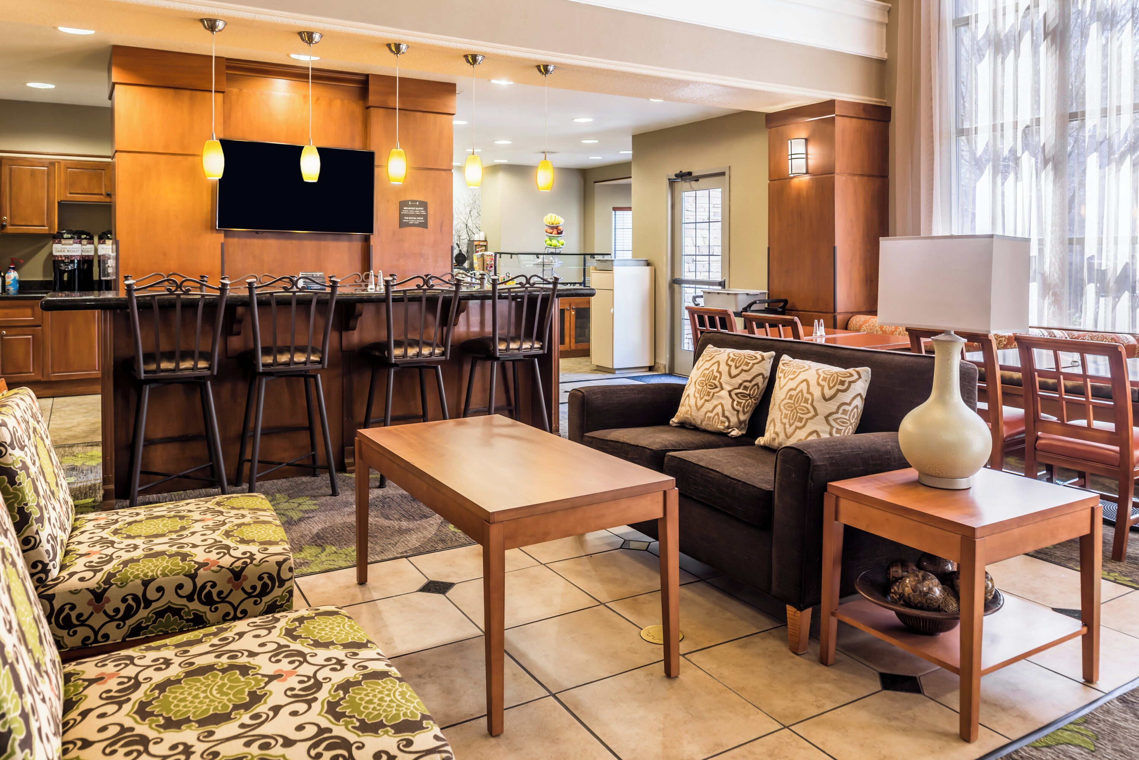 A modern living room with a sofa, armchair, and coffee table. There is a kitchen area with a refrigerator and a flat-screen TV.