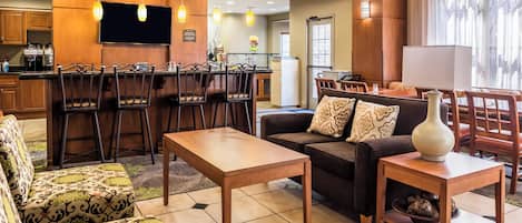 A modern living room with a sofa, armchair, and coffee table. There is a kitchen area with a refrigerator and a flat-screen TV.