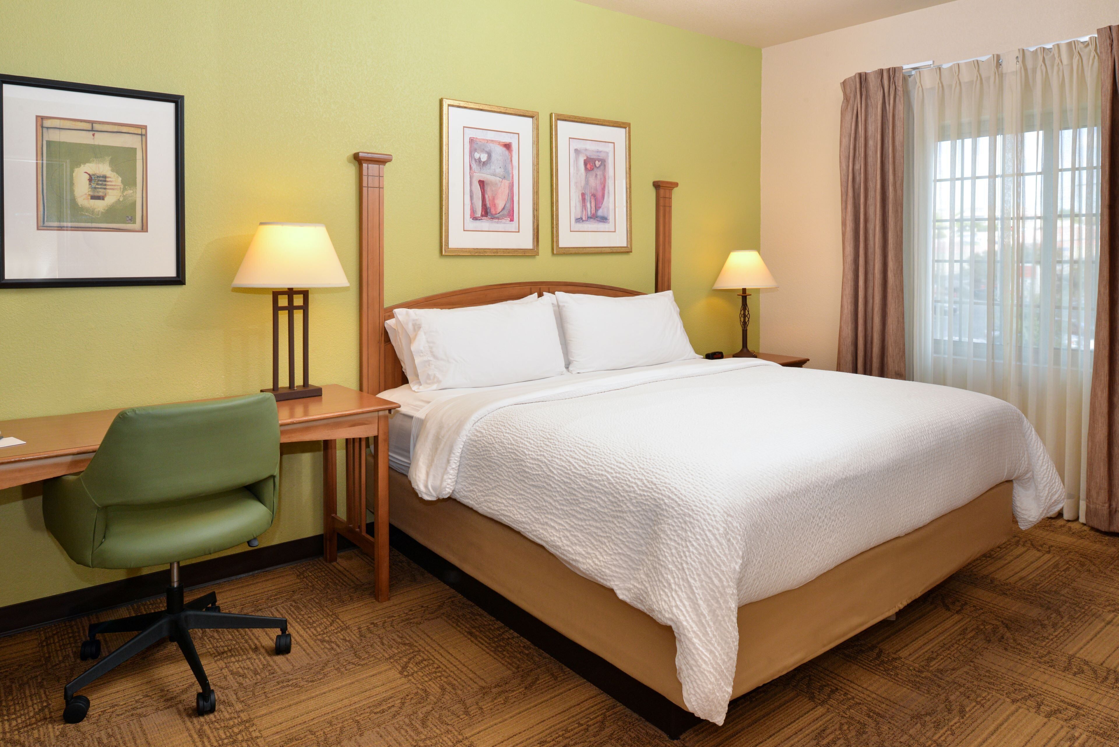 A hotel room with a bed, desk, chair, two lamps, and framed artwork on the wall.