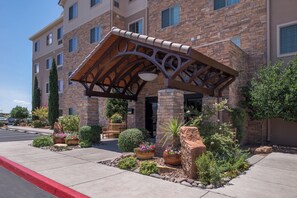 A hotel entrance with a covered patio, stone facade, and landscaped garden with potted plants and a bench.