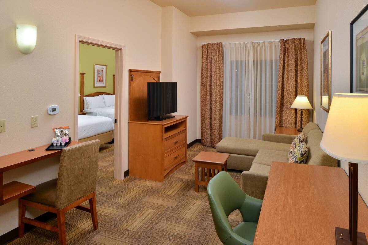 A hotel room with a bed, a TV, a desk, a chair, a sofa, a lamp, and a window with curtains.