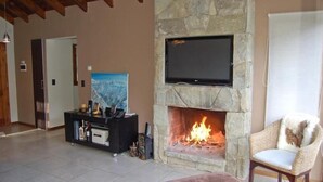 Een woonkamer met een stenen openhaard, een flatscreen televisie erboven en een zwarte tv-kast, plus een rieten fauteuil.