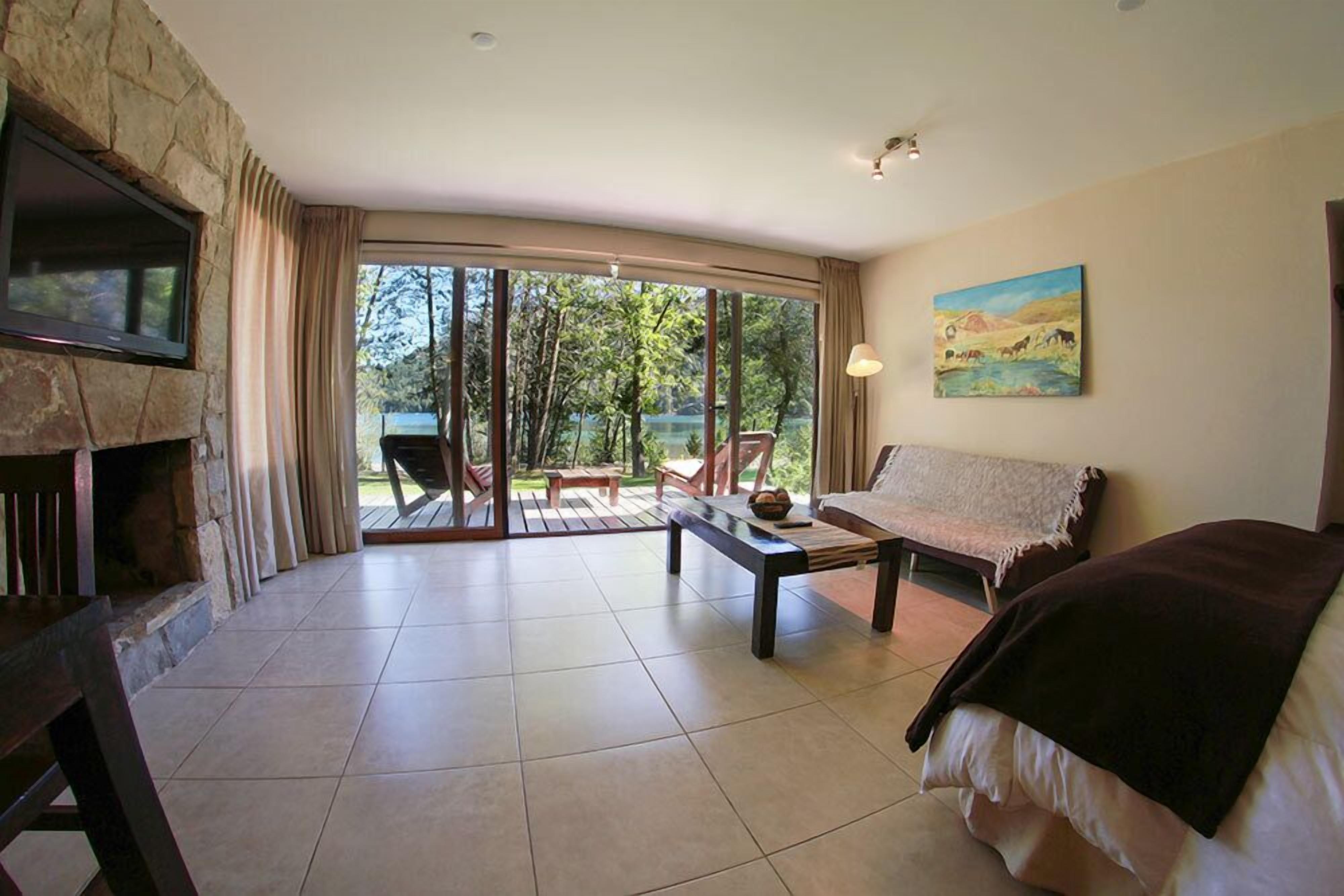 A spacious room with a stone fireplace, a bed, a sofa, and a dining table. There is a sliding glass door leading to an outdoor area with a view of trees and a mountain.