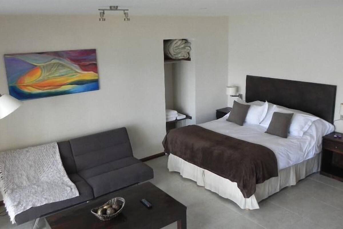 Une chambre d’hôtel avec un lit, un canapé gris, une table basse, un tableau et une table de chevet.