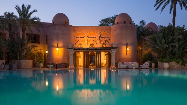 A poolside view of a building with a reflective pool, palm trees, and warm lighting. - Palais Mehdi (Marrakech)