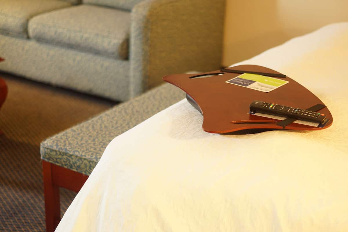 A hotel room with a bed, a remote control, and a small table with a patterned cushion.