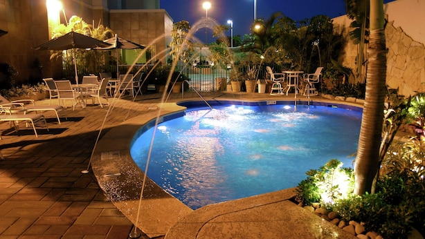 Una piscina con sillas de descanso y sombrillas, rodeada de plantas y un muro de piedra.
