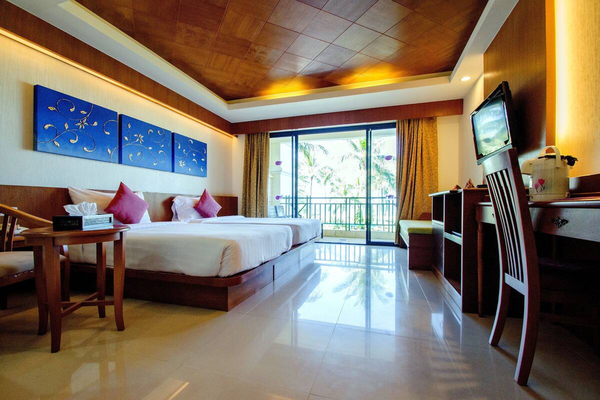 Habitación de hotel con una cama grande, un escritorio, televisión y balcón con vistas a palmeras.