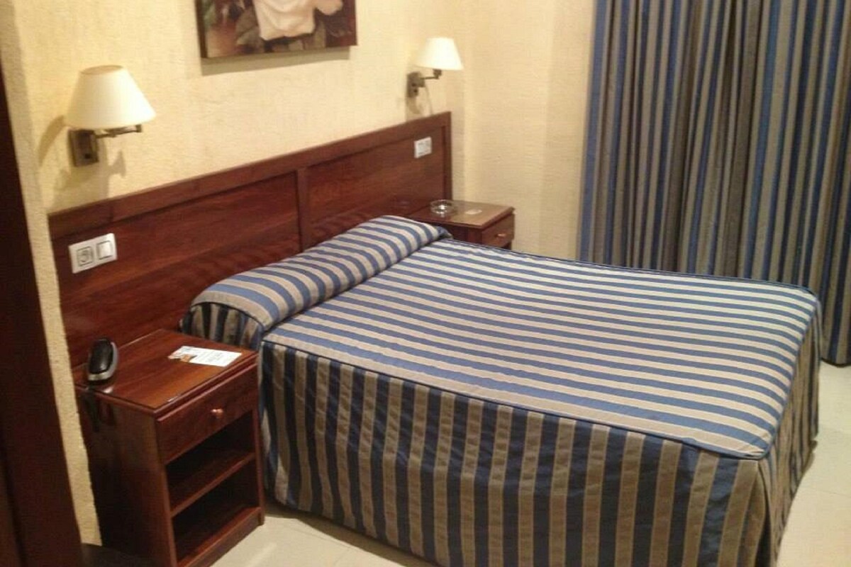 Une chambre d’hôtel avec une tête de lit en bois, une couvre-lit à rayures, deux lampes de chevet, une table de chevet, un téléphone et un tableau représentant des roses accroché au mur.