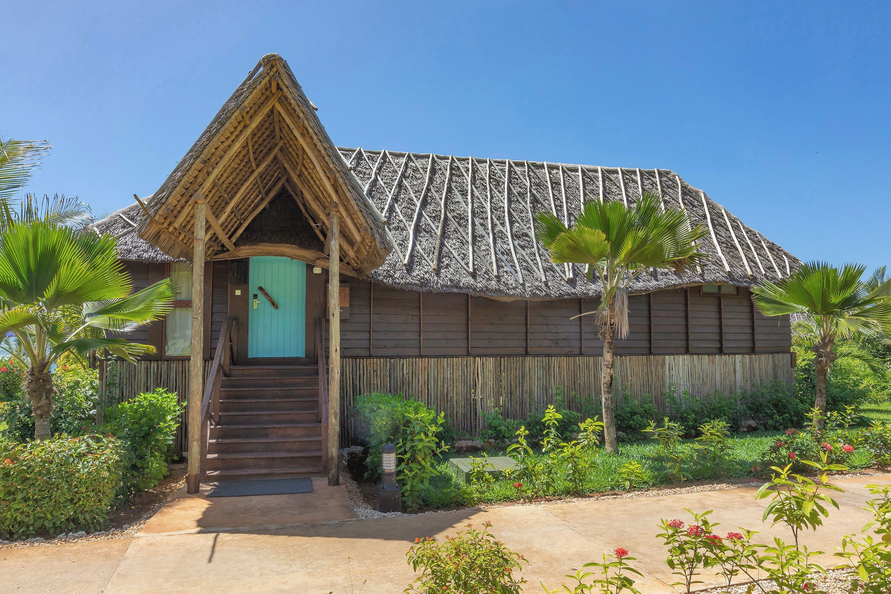 A traditional thatched-roof building with a wooden staircase leading to a door, surrounded by lush greenery and a clear sky.