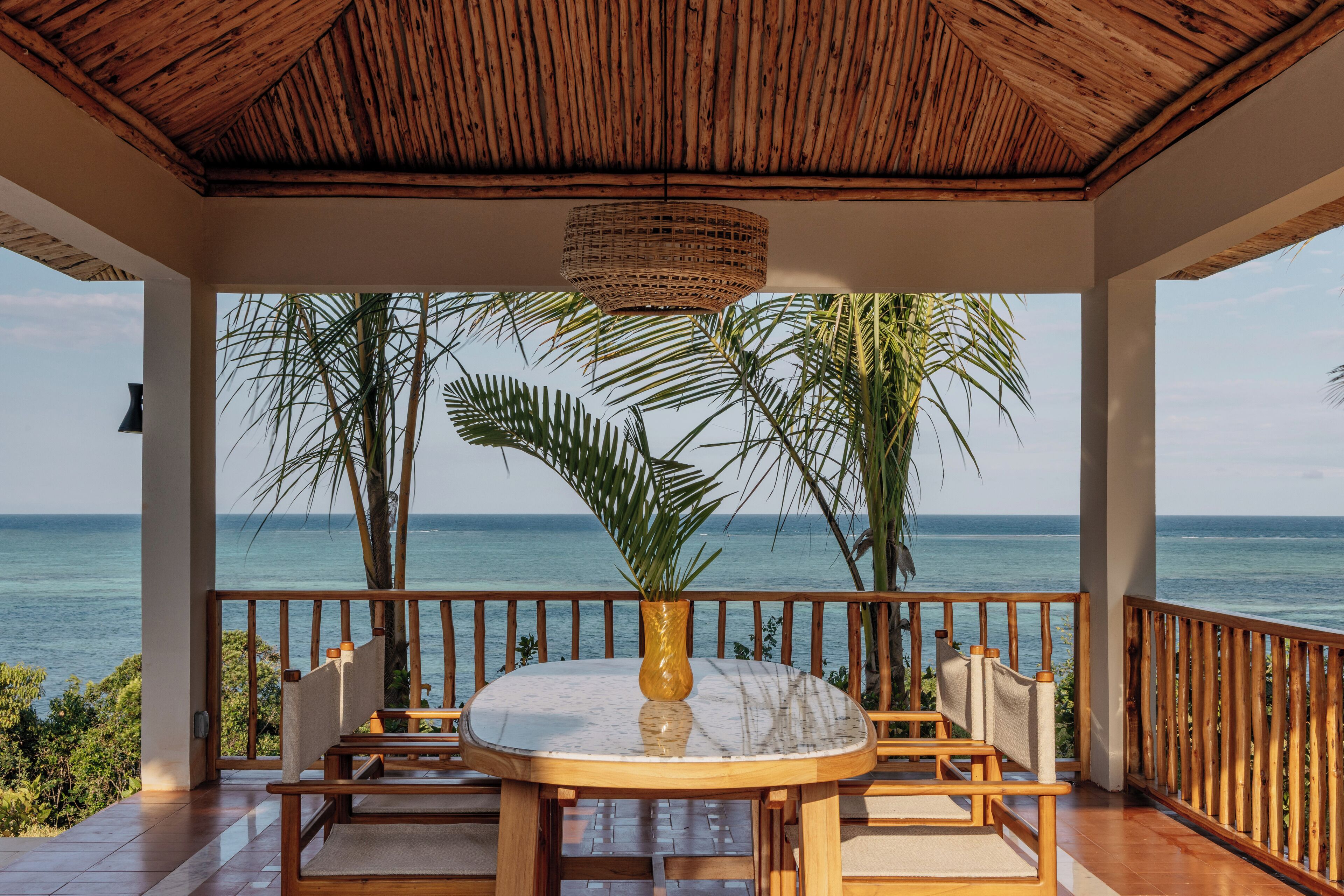A covered outdoor seating area with a table and chairs, overlooking a beach with palm trees and the ocean.