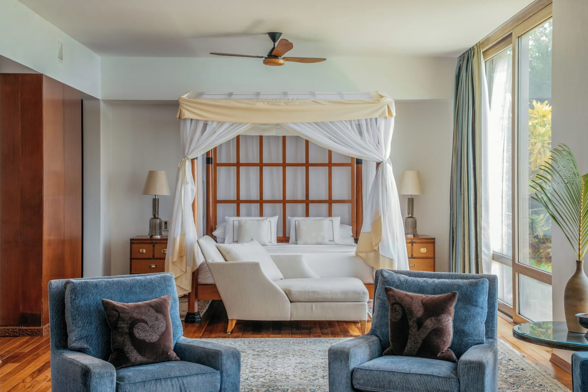 Ein Schlafzimmer mit einem Baldachinbett, zwei Sesseln, einem Nachttisch mit Lampe und einem großen Fenster mit Vorhängen.