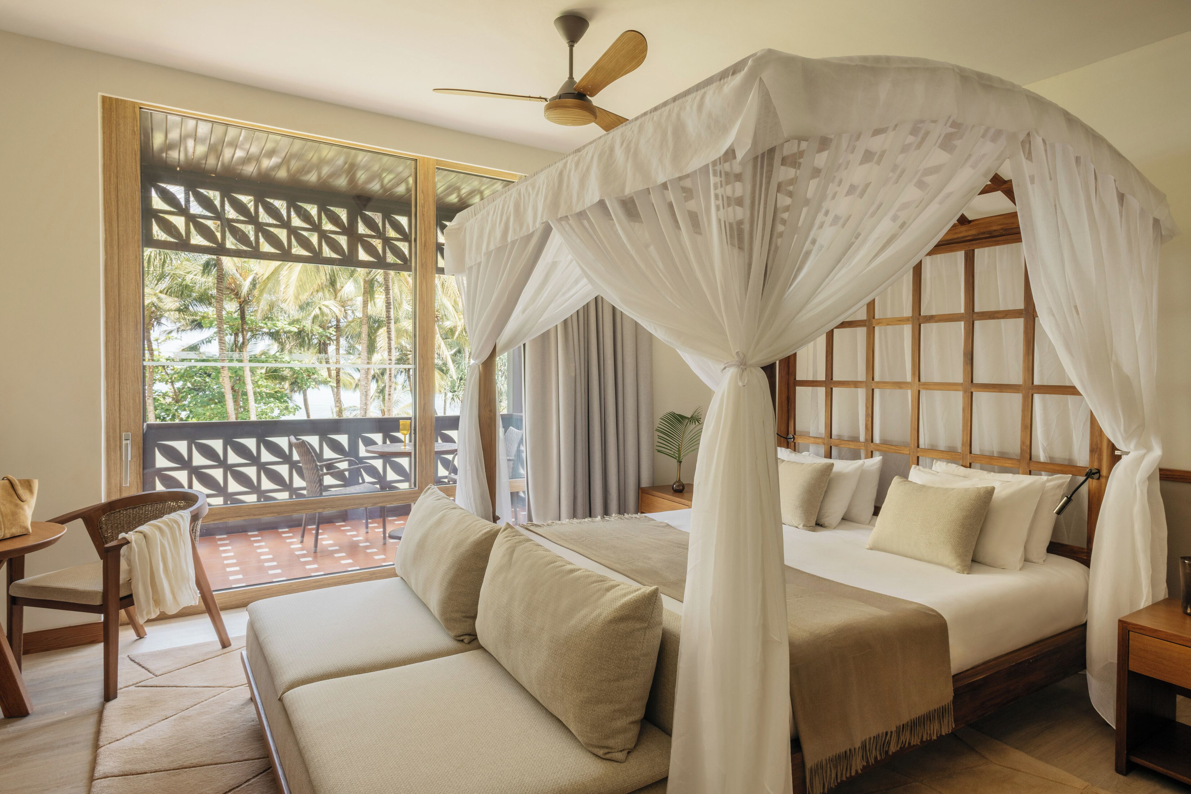 A bedroom with a bed, a sofa, a ceiling fan, and a view of palm trees through a window.