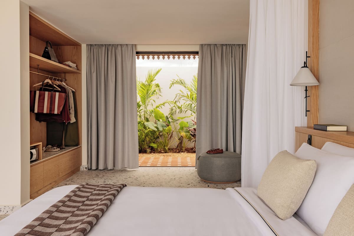 Ein Schlafzimmer mit Bett, Kleiderschrank, Nachttisch mit Lampe und Blick auf einen Garten durch ein fenster mit Vorhängen.