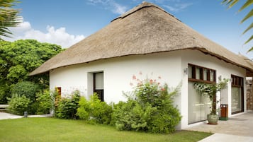 A thatched-roof house with a white exterior, surrounded by lush greenery and a clear sky.