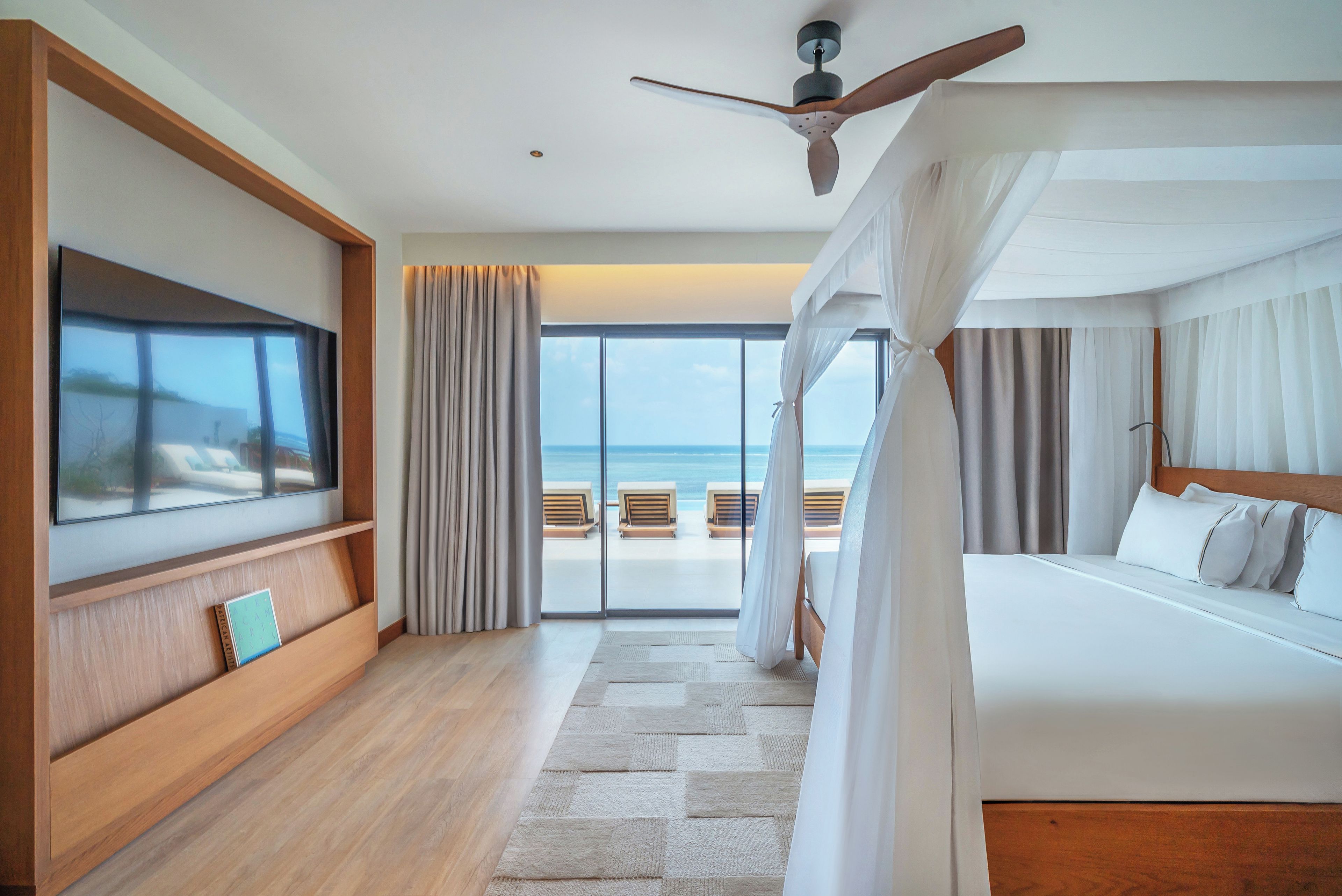 A hotel room with a large TV, a ceiling fan, a bed with white bedding, a view of the beach, and a balcony with lounge chairs.